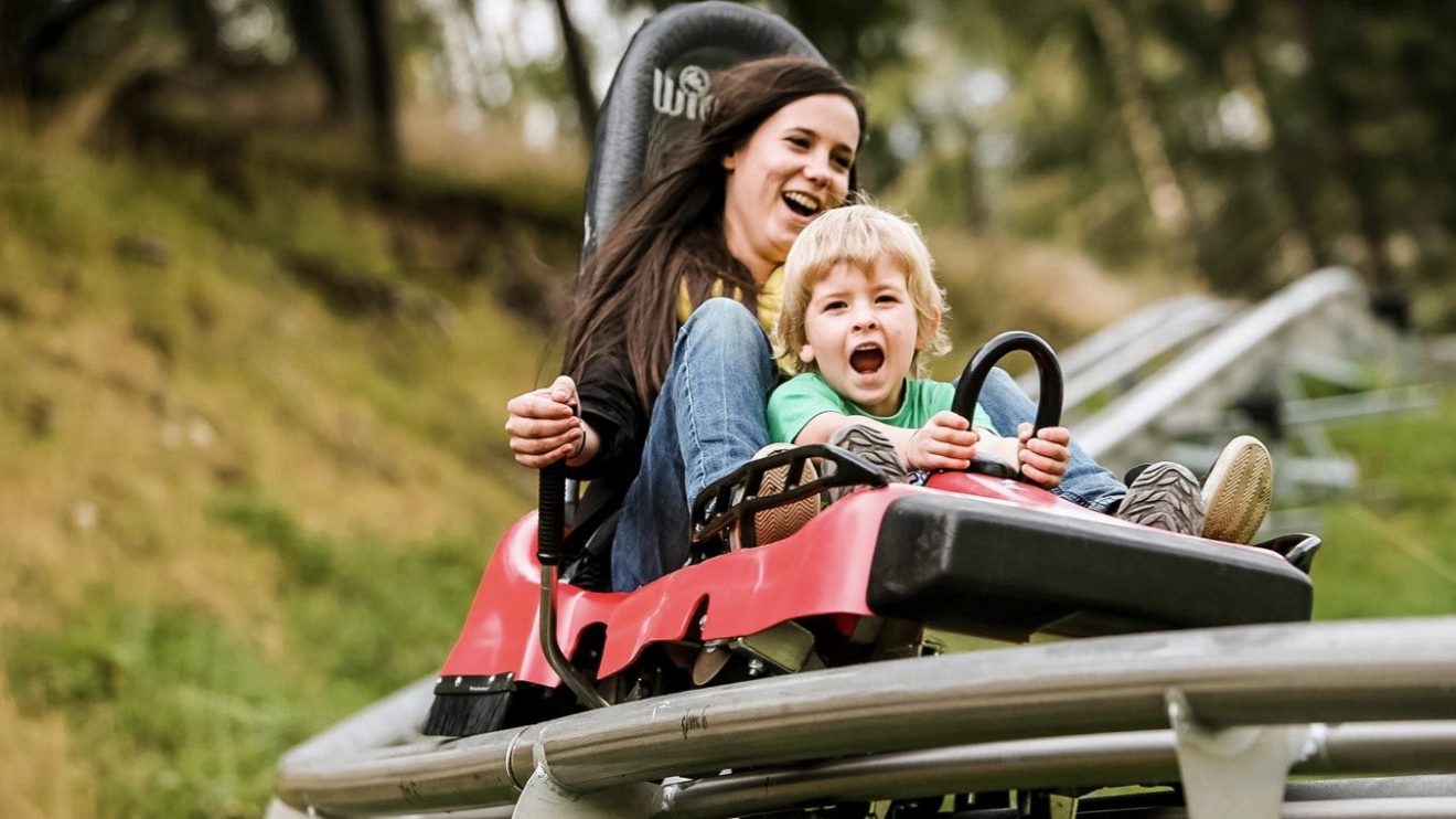 Woman and child joyfully riding a red sled on a track in a grassy area.