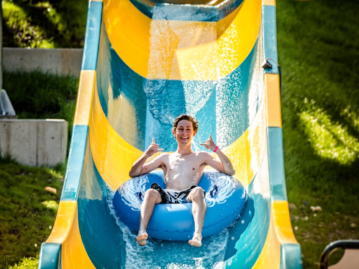 Person on yellow and blue water slide with inner tube, giving thumbs up, sunny day.