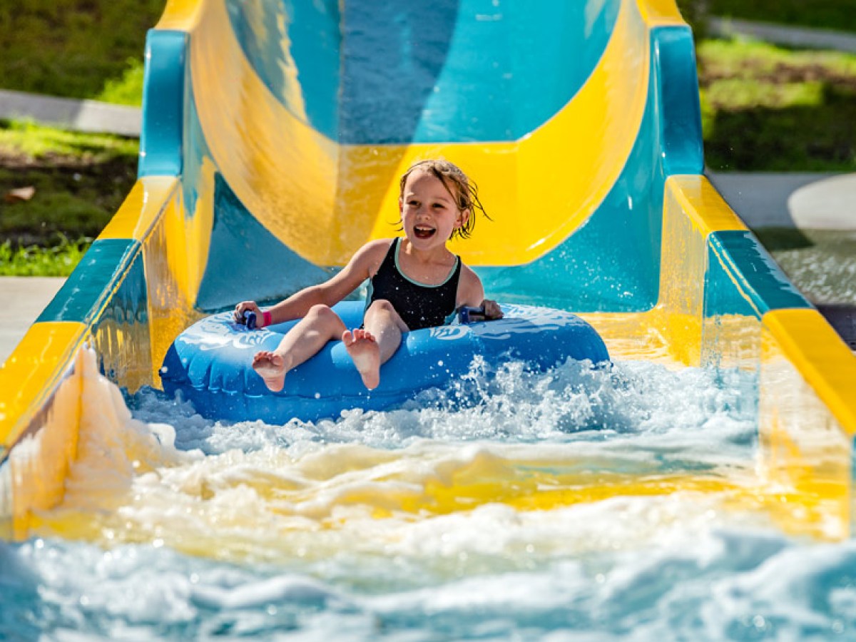 Child on a blue inflatable tube sliding down a yellow and blue waterslide, smiling.