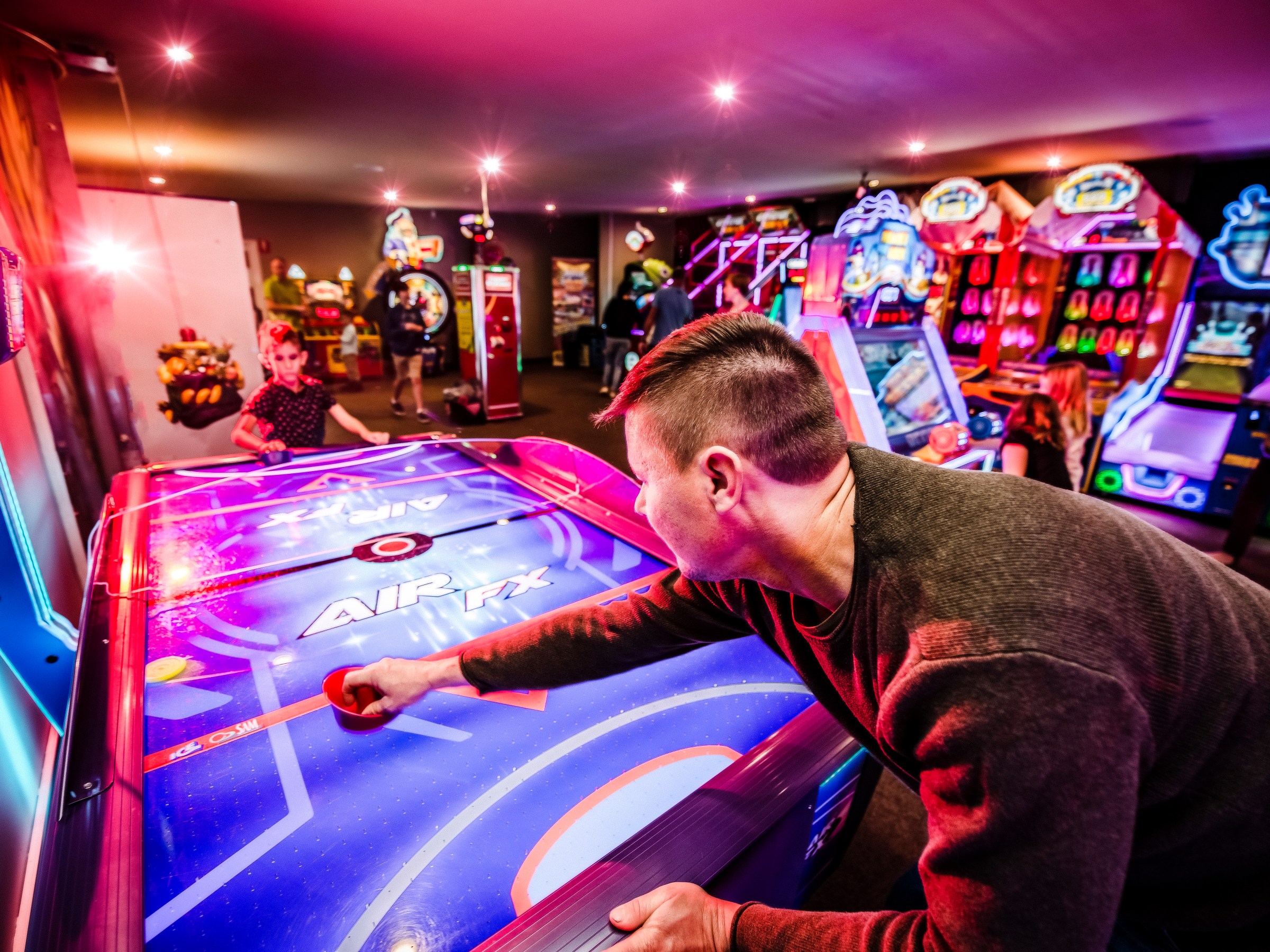 Man playing air hockey in a colorful arcade with bright lights and various games in the background.
