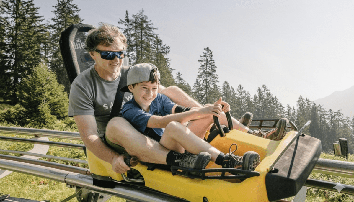 Two people smiling on a yellow alpine coaster with scenic forest in the background.