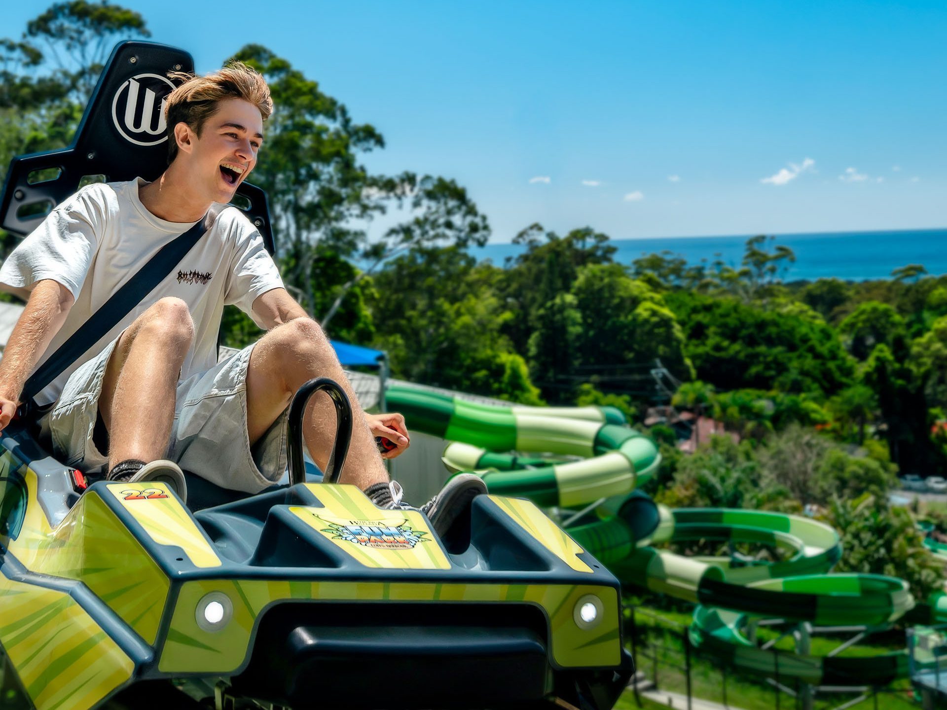 A person riding a roller coaster with scenic views of trees and ocean in the background under a clear blue sky.