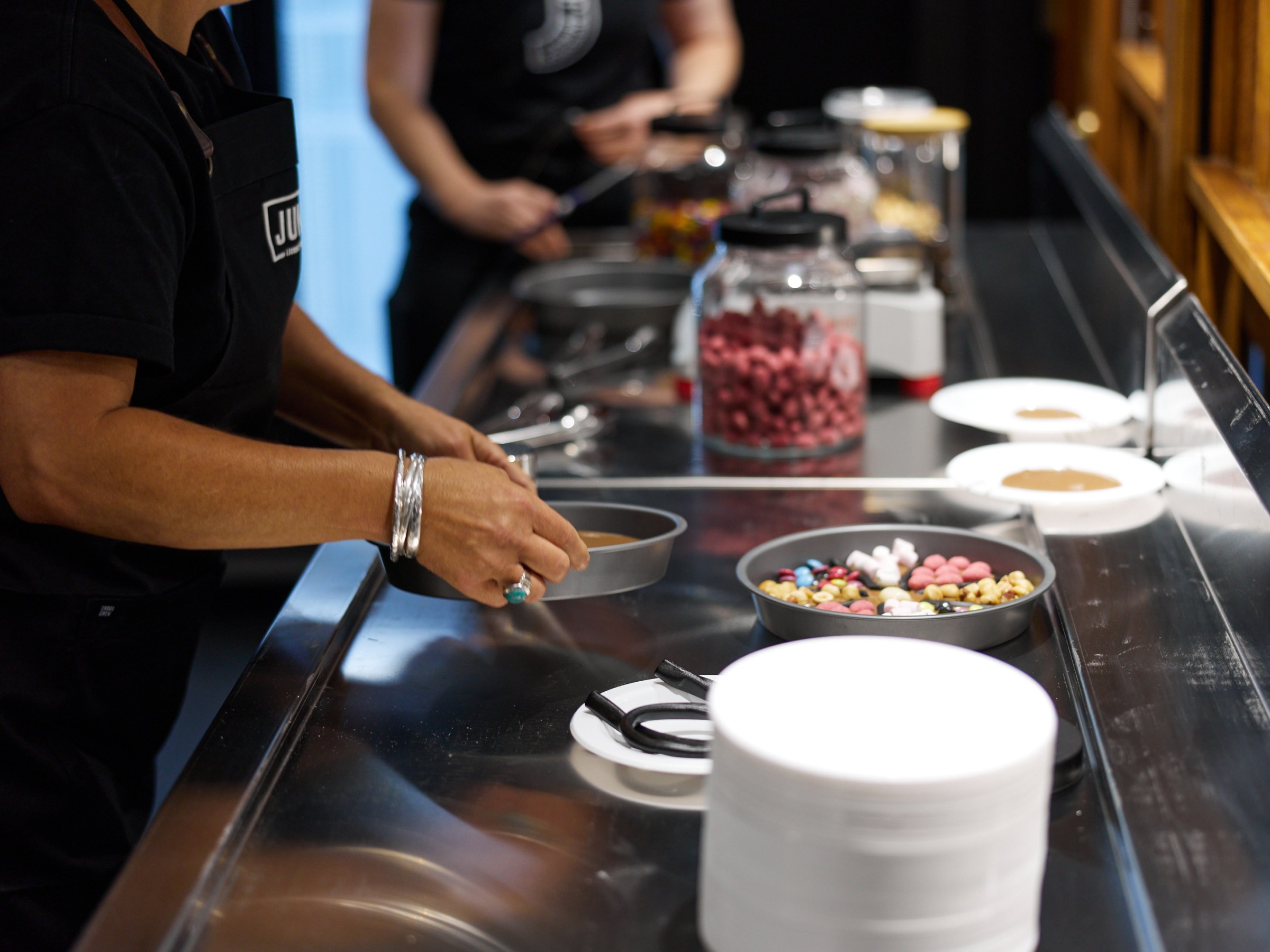 Person wearing black apron preparing toppings at a counter with jars and trays.