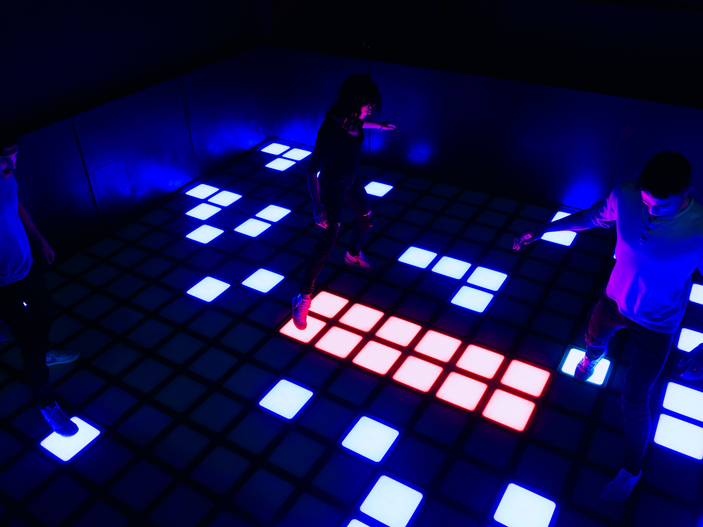 People playing on an interactive floor with illuminated blue and red squares in a dark room.