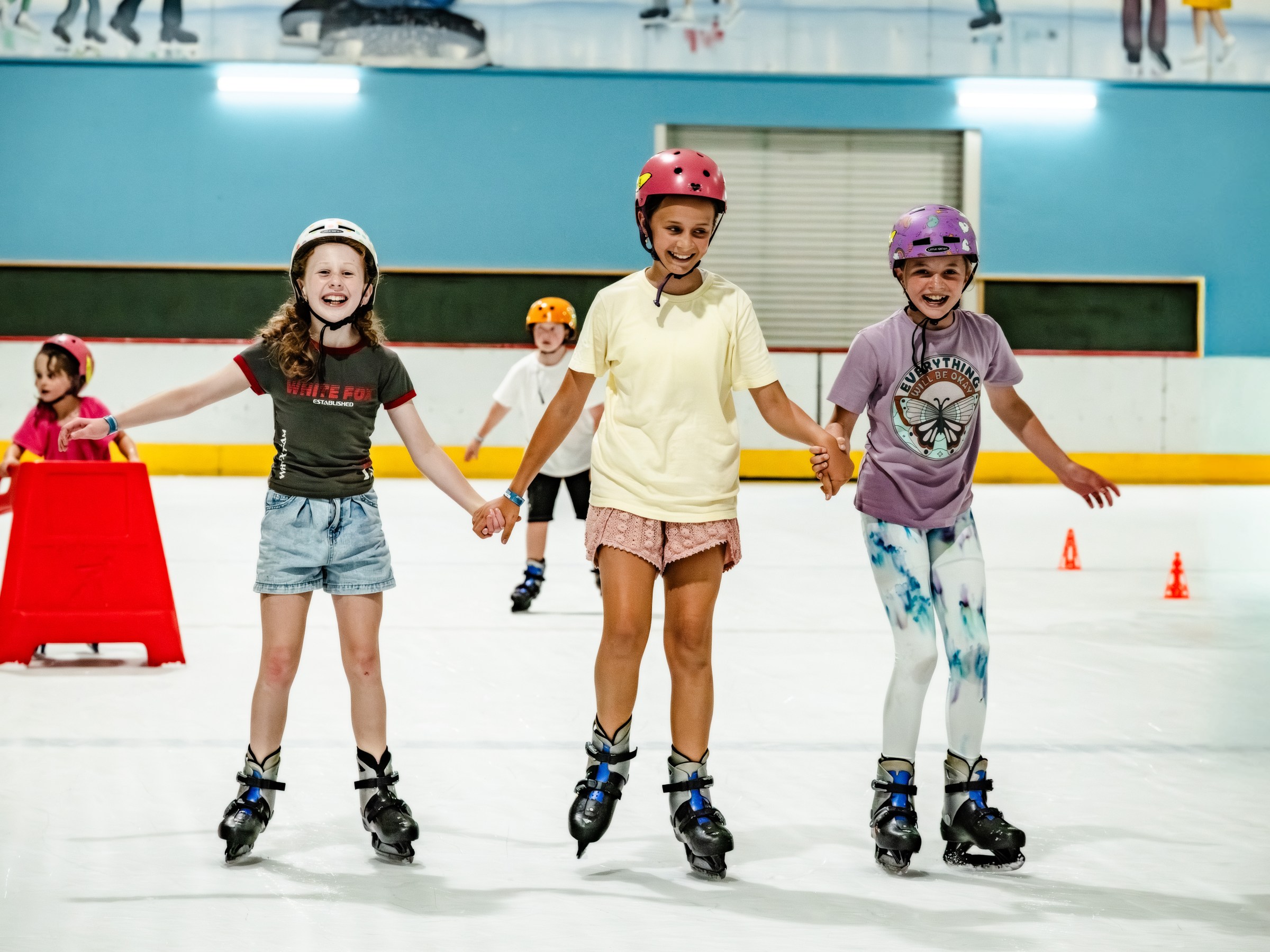 Three kids in helmets holding hands while ice skating indoors.