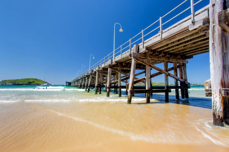 Sunny beach with wooden pier extending over clear water and blue sky.