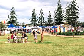 People gathering at an outdoor market with tents and tall pine trees.