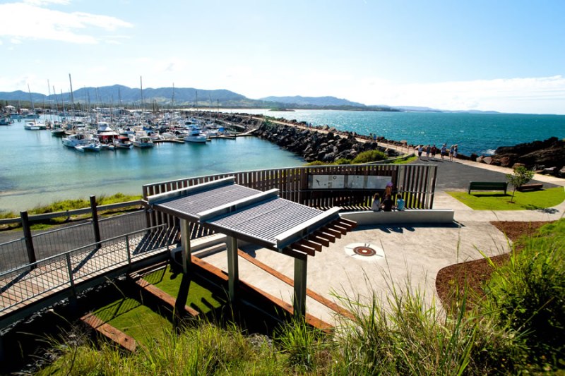 Coastal marina with boats, walkway, and grassy landscape under a clear blue sky.