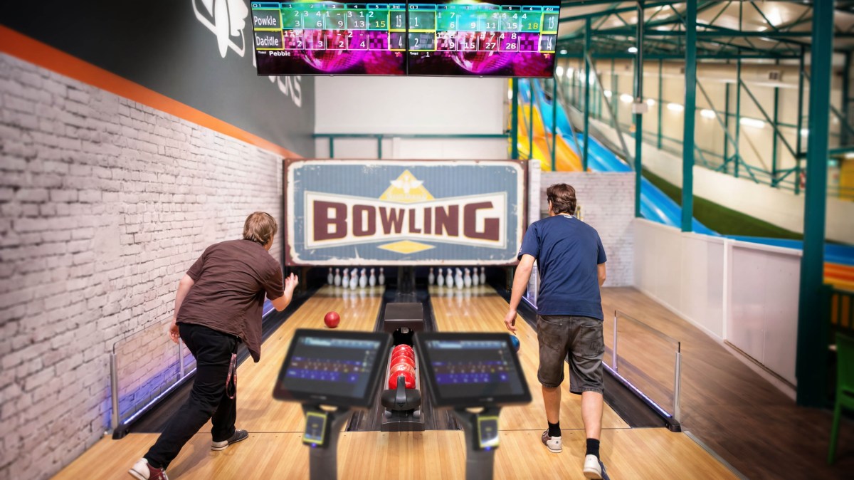 Two people bowling indoors with electronic scoreboards overhead.