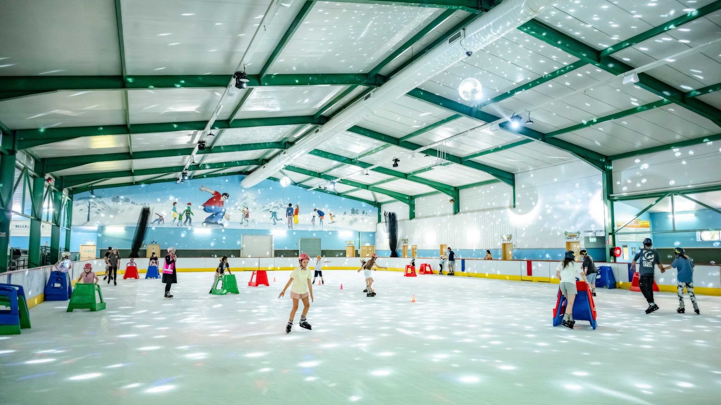 Indoor ice rink with skaters, colorful lights, and a mural on the wall.