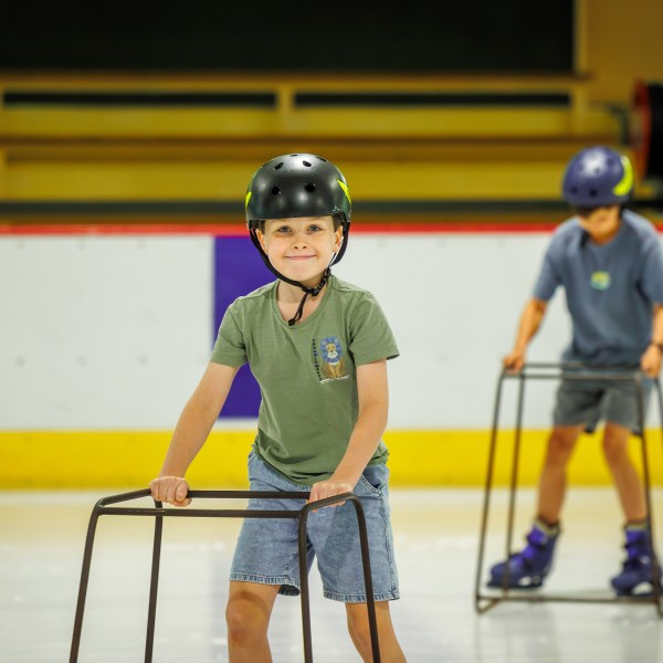 Two children ice skating with helmets and balance supports.