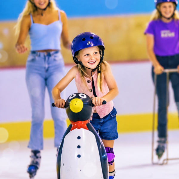 Child with helmet skating, holding penguin support on ice rink.