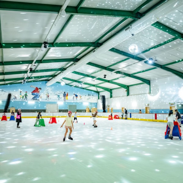 People ice skating indoors under sparkling lights with supporting aids.