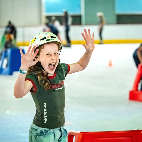 Excited child in helmet ice skating indoors, arms raised.