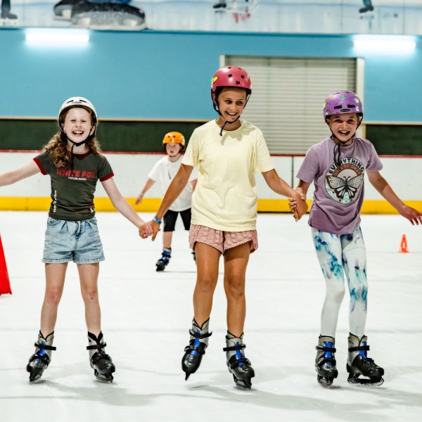 Three kids holding hands roller skating indoors, wearing helmets.